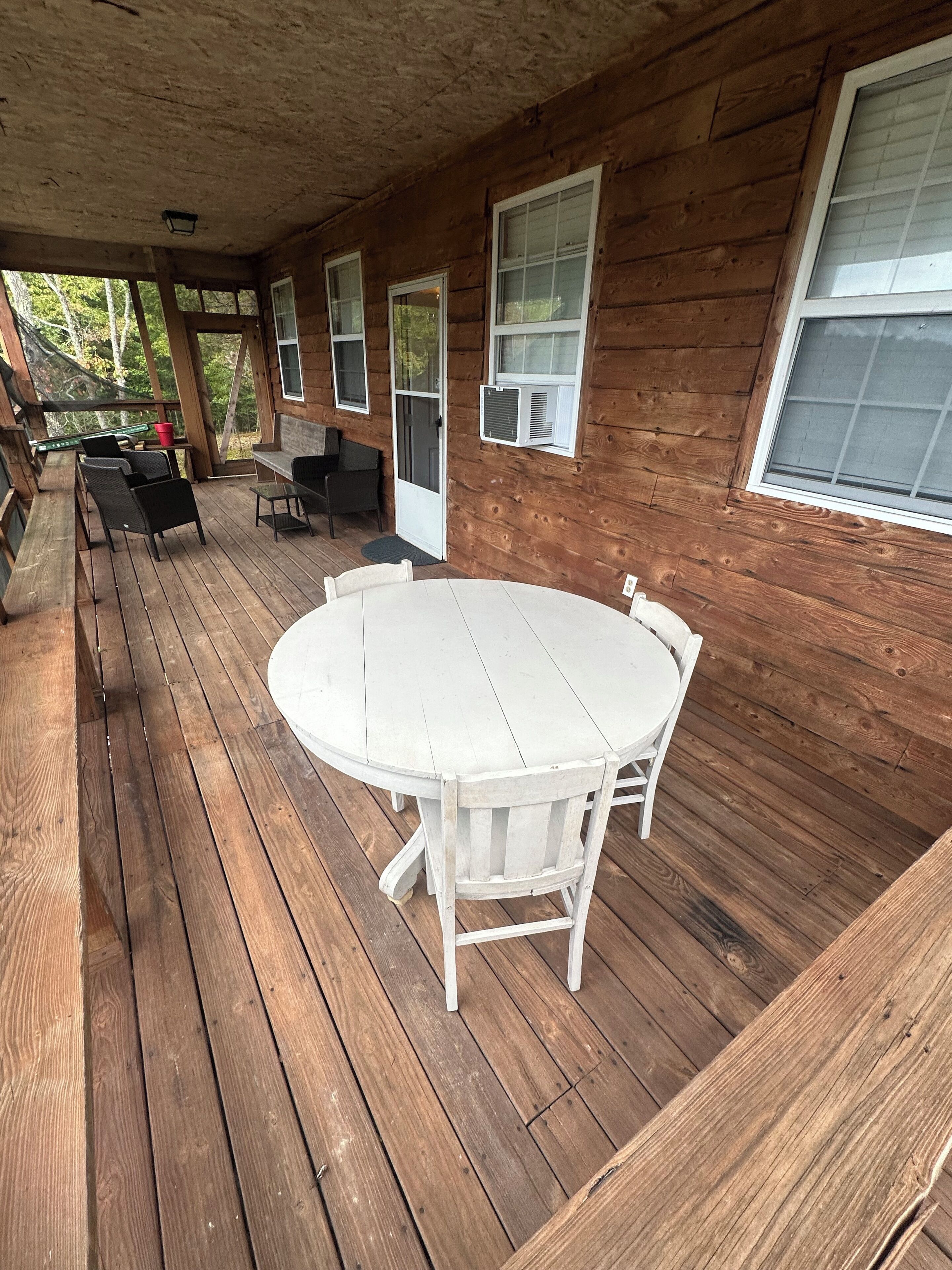 A wooden deck with a round table and chairs, a covered seating area with chairs, and a window with an air conditioning unit.