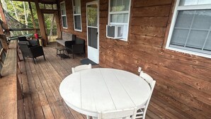 A wooden deck with a round table and chairs, a covered seating area with chairs, and a window with an air conditioning unit.