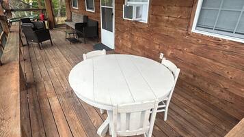 A wooden deck with a round table and chairs, a covered seating area with chairs, and a window with an air conditioning unit.