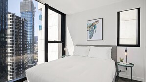 A modern hotel room with a large bed, a city view, a framed artwork, and a small table with a lamp. - Stylish 2-bed Apartment With Stunning City Views (Melbourne)