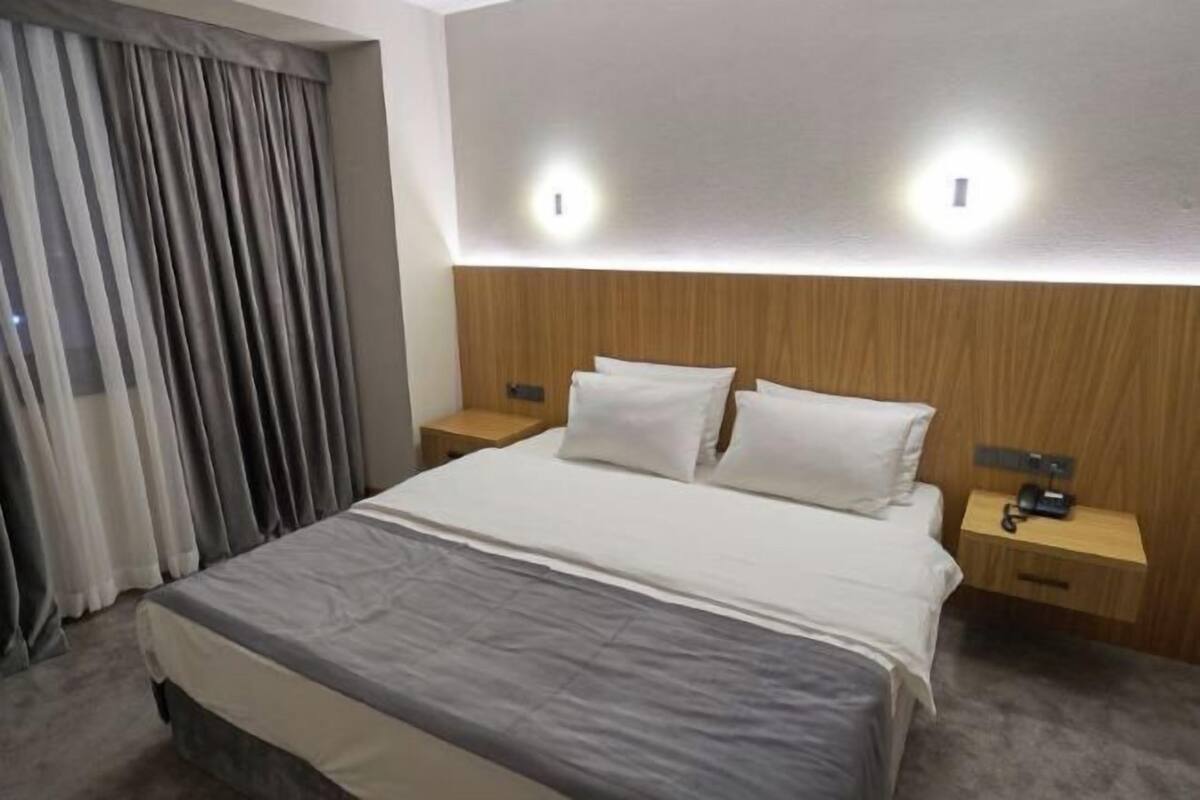 A hotel room with a bed, two bedside tables, a wooden headboard, and a window with curtains.