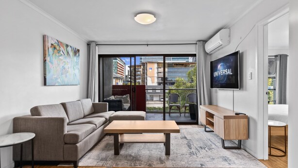 A modern living room with a grey sofa, a wooden coffee table, a TV mounted on the wall, and a view of the city through large glass doors. - Central Bowen Hills Stay (Brisbane)