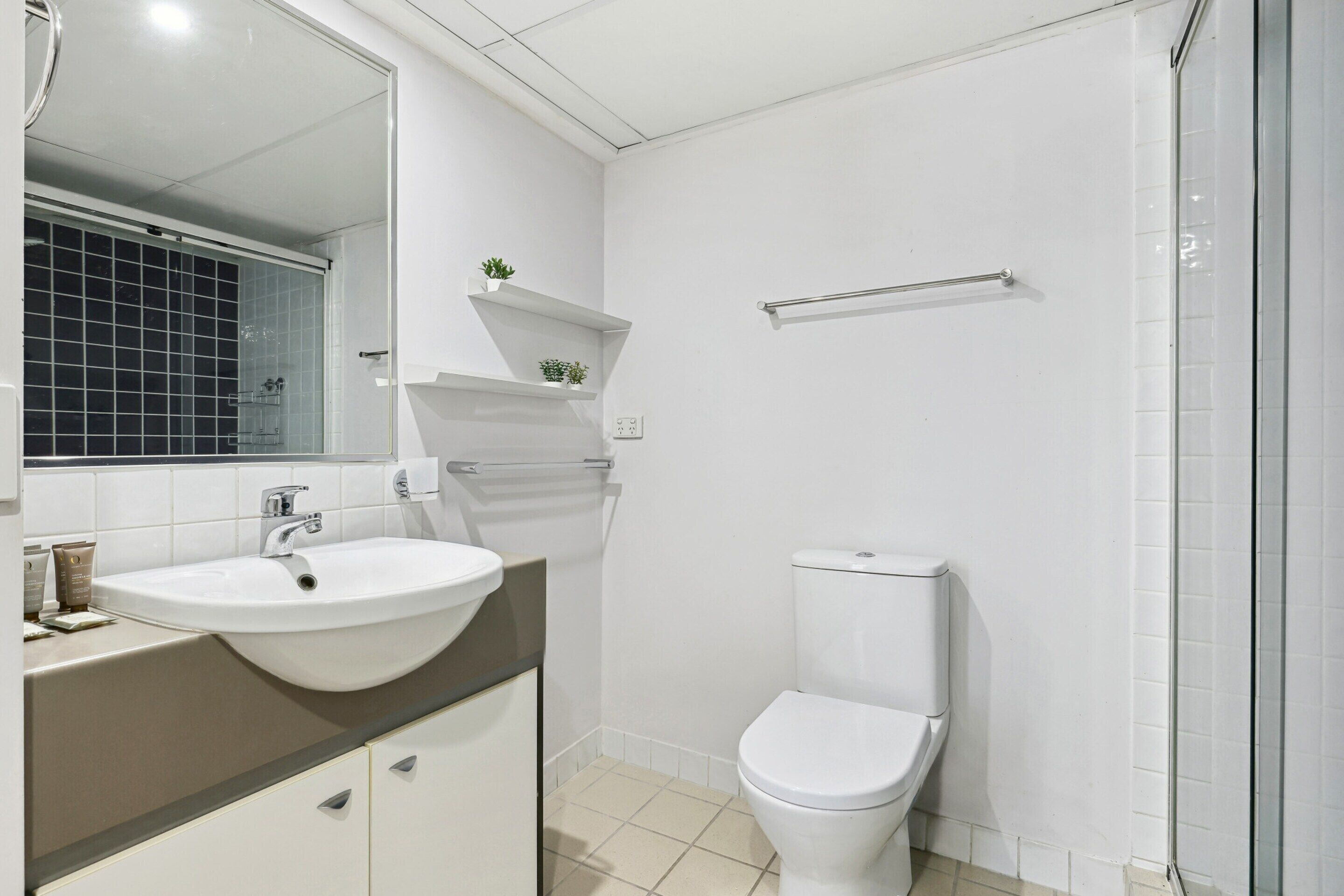 Ein modernes Badezimmer mit einem weißen Waschbecken, einem Spiegel und einer Toilette. Es gibt Regale mit Pflanzen und einen Handtuchhalter.