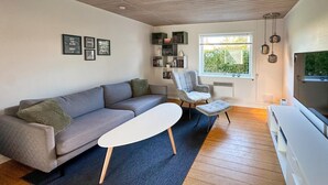 A modern living room with a grey sofa, a white coffee table, and a grey armchair. There is a bookshelf with books and decorative items, a window with a view of greenery, and a ceiling with wooden beams. - 2 bedroom gorgeous home in østermarie (Østermarie)