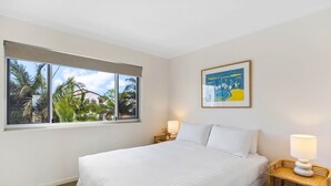 A hotel room with a large bed, a window with a view of palm trees and buildings, a framed picture on the wall, and two bedside lamps. - Oceanfront 3-bed Retreat With Pool-steps to Beach (Sunshine Coast)