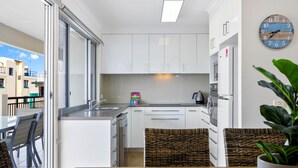 A modern kitchen with white cabinets, a wooden dining table, wicker chairs, and a potted plant. - Oceanfront 3-bed Retreat With Pool-steps to Beach (Sunshine Coast)