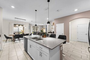 A modern kitchen with a central island, pendant lighting, and a dining area with chairs and a table.