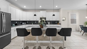 A modern kitchen with white cabinets, a stainless steel refrigerator, a central island with bar stools, pendant lighting, and a dining area with a table and chairs. - DisneyVilla 6 BD, Club/Water Park/Pool/Spa and BBQ (Kissimmee)