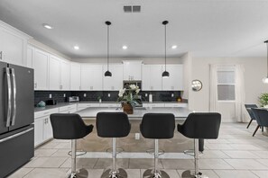 A modern kitchen with white cabinets, a stainless steel refrigerator, a central island with bar stools, pendant lighting, and a dining area with a table and chairs.