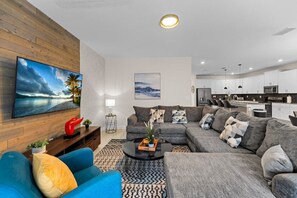 A modern living room with a sofa, coffee table, and TV. There is a kitchen in the background.