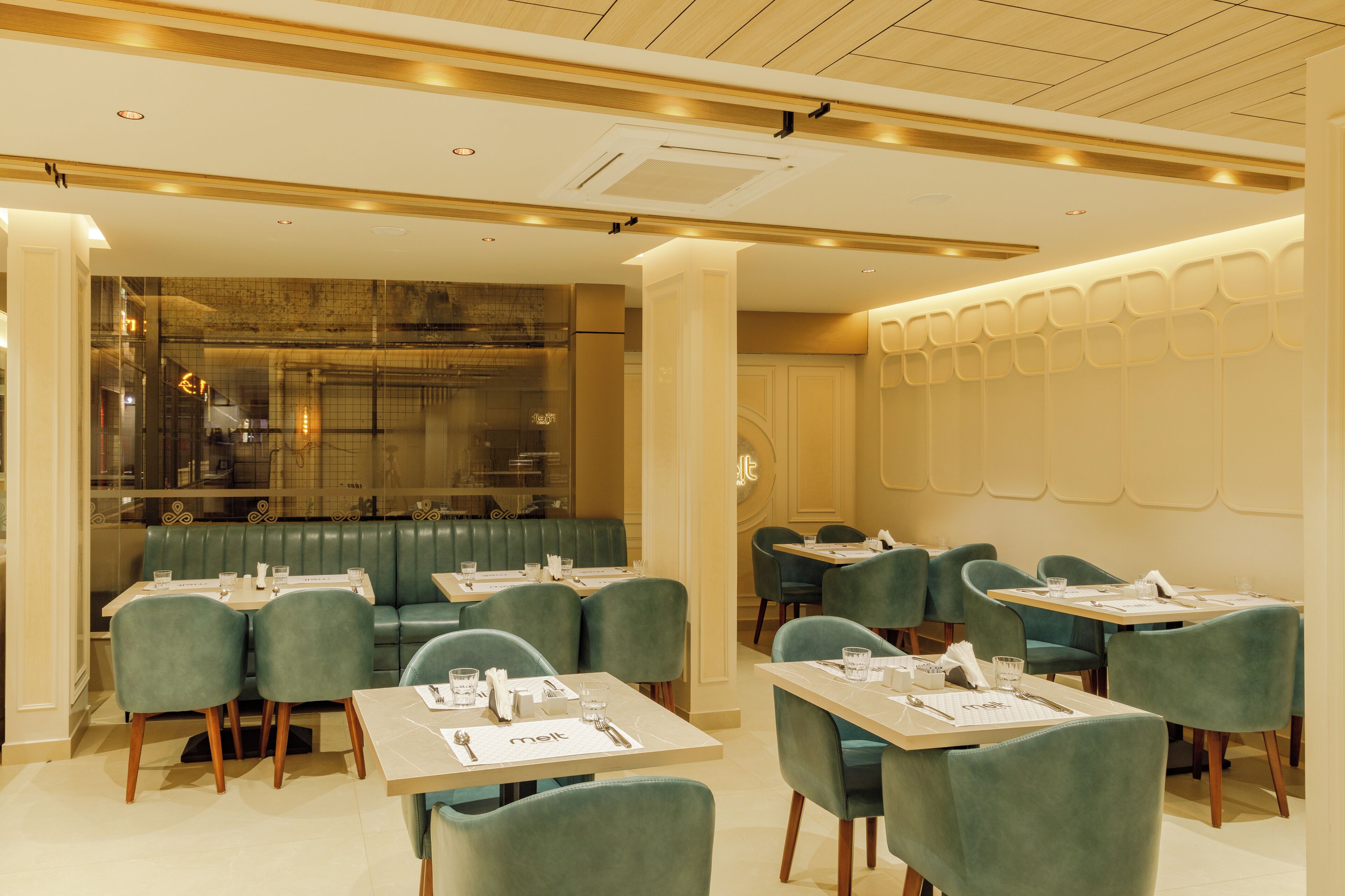 A restaurant with green upholstered booths and tables, a modern ceiling design, and a glass partition separating the dining area from the kitchen.