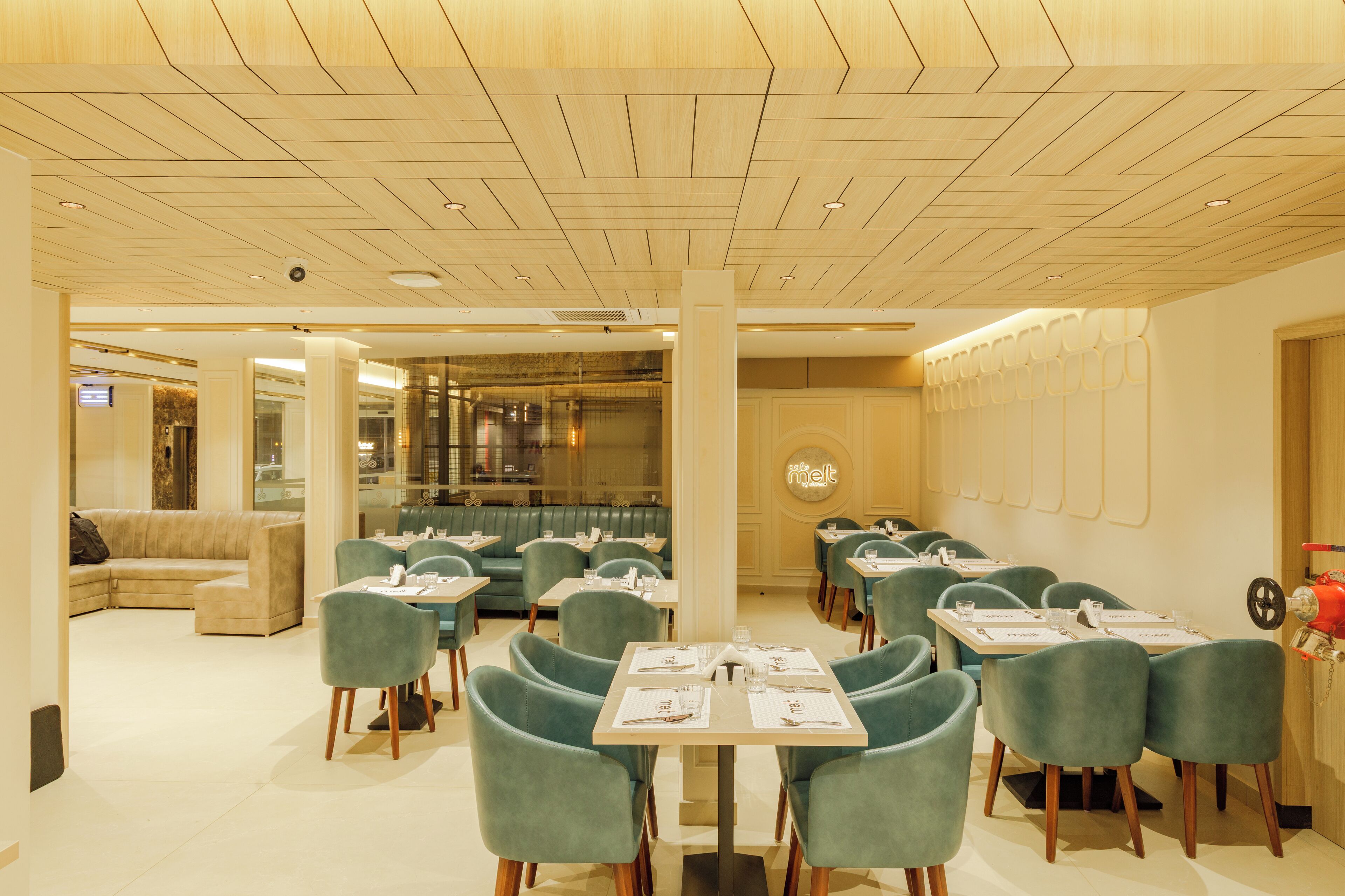 A modern dining area with green upholstered chairs and tables, a wooden ceiling, and a glass partition separating the dining area from a seating area.