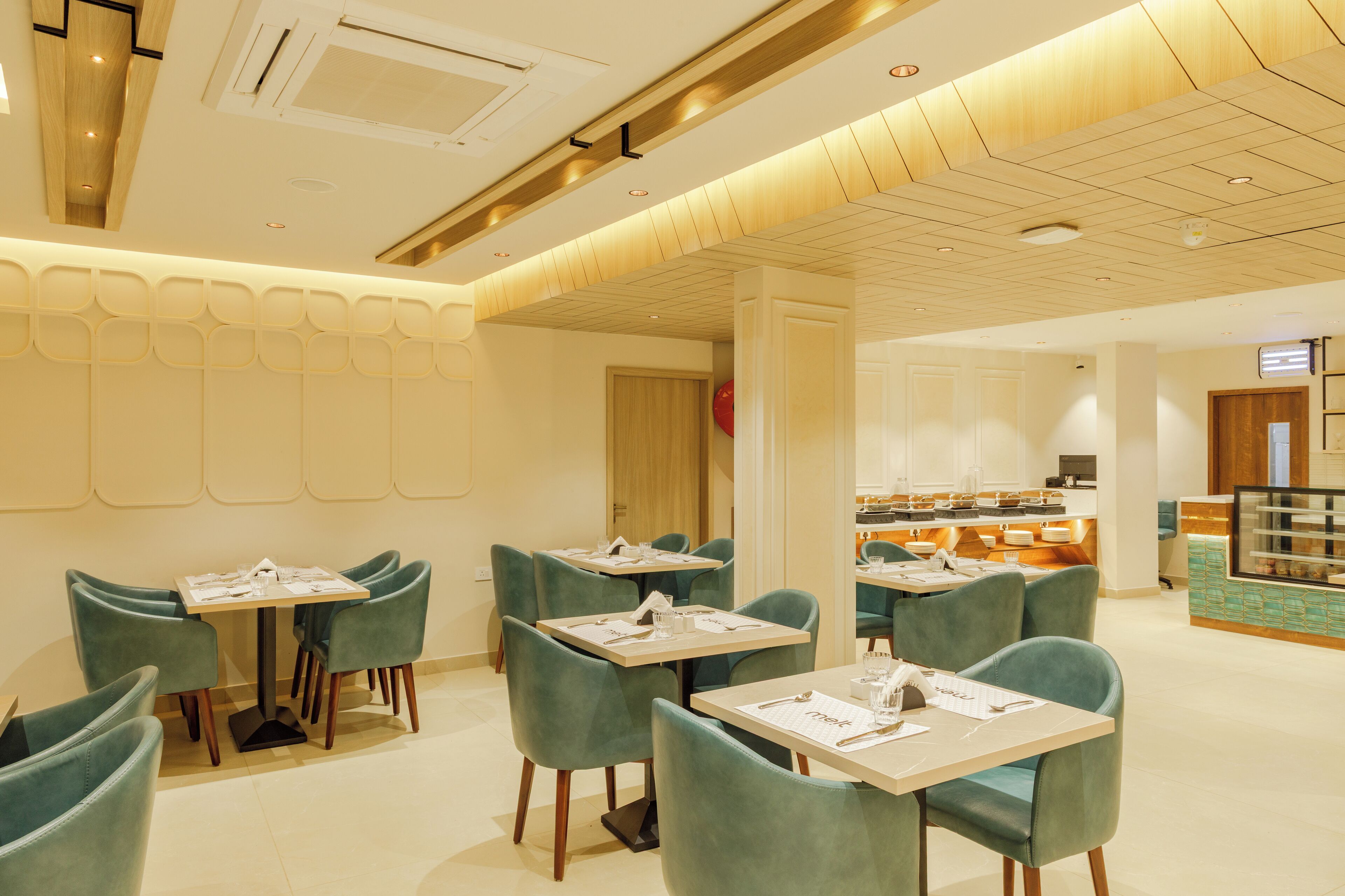 A modern dining area with teal chairs and tables, a kitchen in the background, and a wooden ceiling.