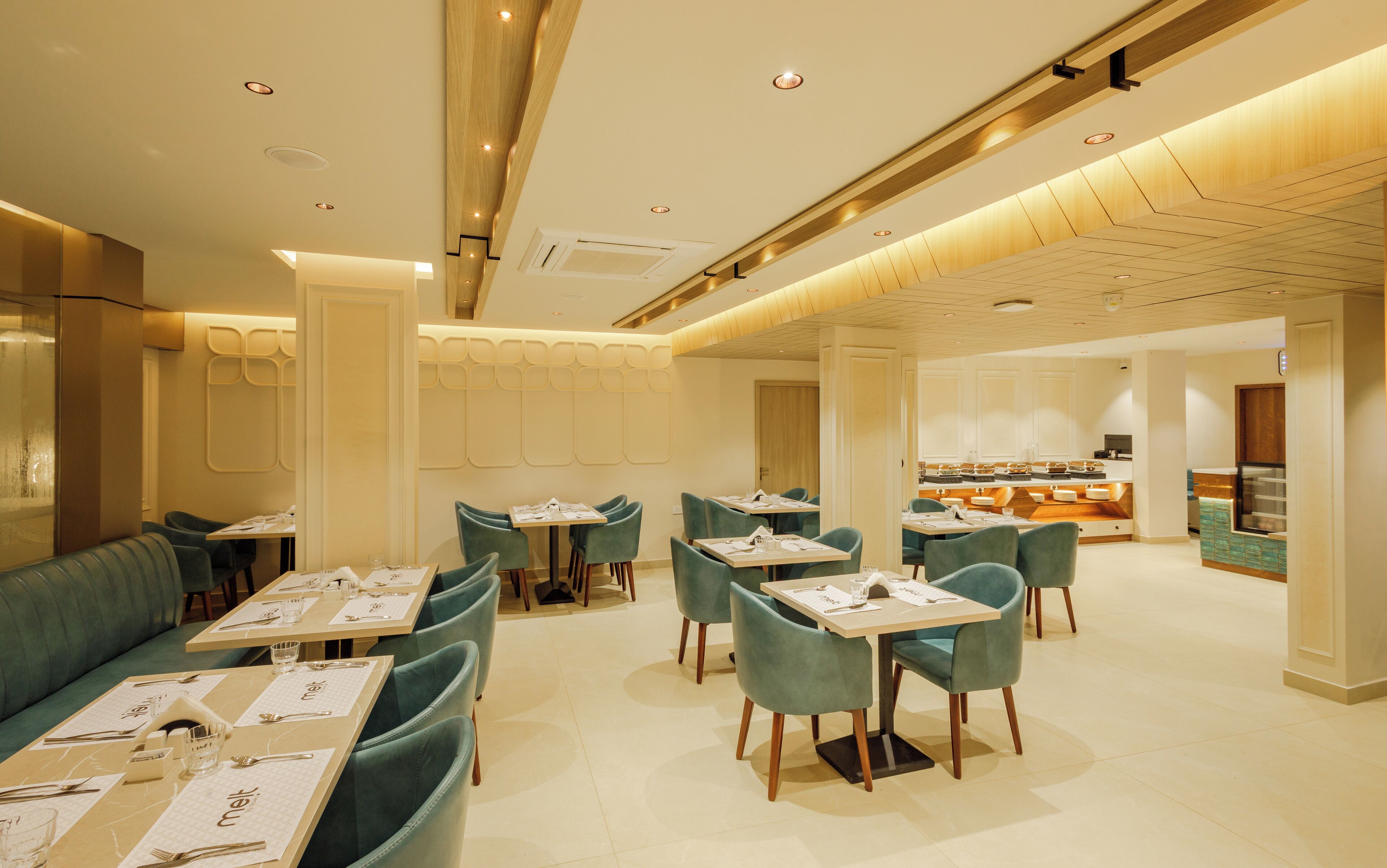 A modern restaurant with teal upholstered booths and tables, a kitchen area in the background, and a warm, well-lit ambiance.