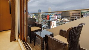 Un balcone con mobili in vimini e un tavolo in vetro, con vista su un panorama urbano che include edifici e una strada.