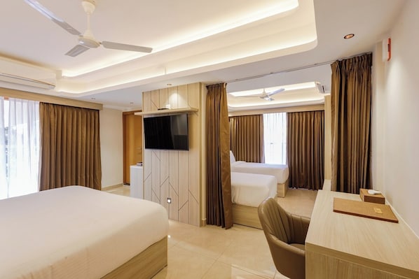 A hotel room with a bed, a desk, a chair, a TV, and a ceiling fan. - Olwen Suites (Kanayannur)