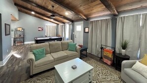 A cozy living room with a wooden ceiling, a beige sofa, a white coffee table, and a pool table.