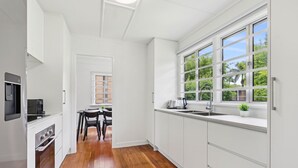 Una cocina moderna con armarios blancos, piso de madera, un ventanal con vistas a la vegetación y un comedor con mesa y sillas.
