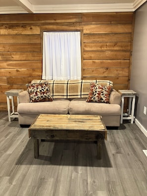 A cozy living room with a beige sofa, two patterned throw pillows, a wooden coffee table, and a window with white curtains. - Cozy 1-bedroom railroad house (Lima)