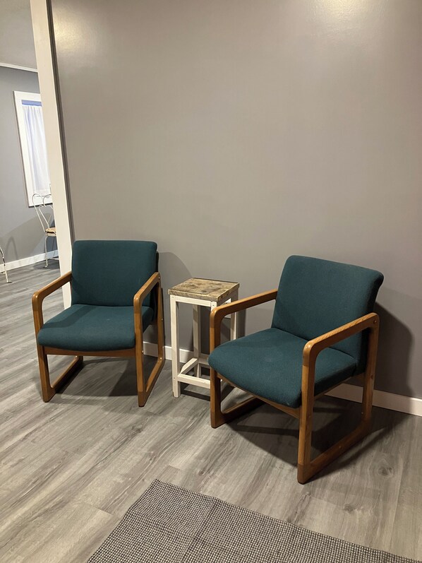 Deux fauteuils turquoise à armature en bois et une petite table d’appoint blanche avec un plateau en bois, placés sur un sol gris, avec un mur gris en arrière-plan.