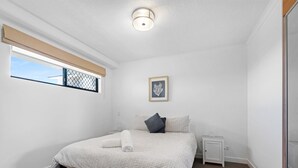 Une chambre à coucher comprenant un lit, une table de chevet, une commode, une fenêtre avec des stores, un luminaire au plafond et un tableau encadré accroché au mur.
