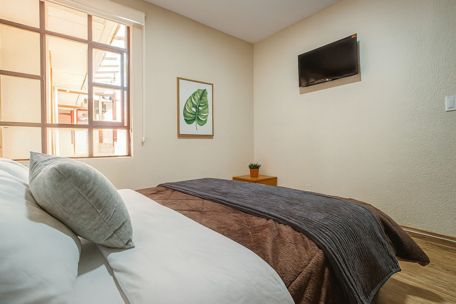 A bedroom with a bed, a window, a wall-mounted TV, and a framed plant.