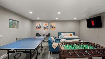 Een kamer met een tafeltennistafel, een tafelvoetbalspel, een bed, een bank en een televisie.