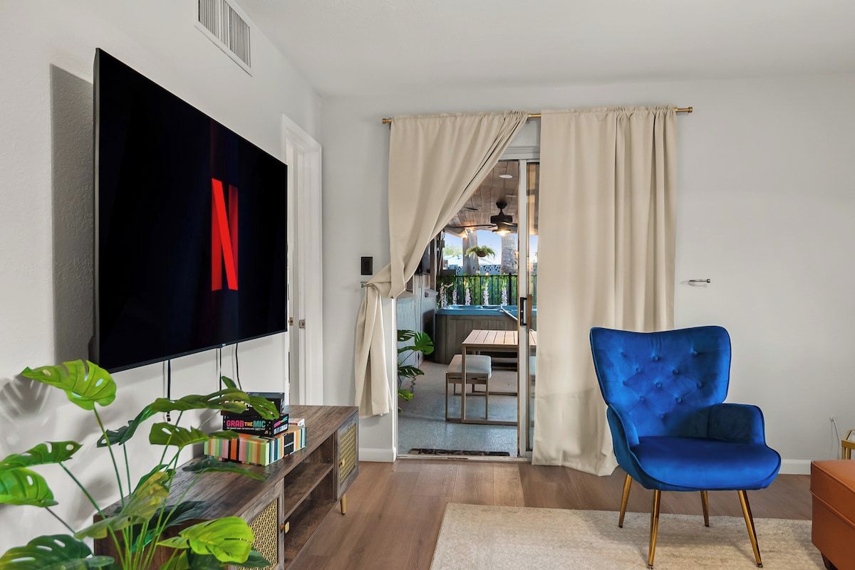 A living room with a flat-screen TV, a blue armchair, a bookshelf, and a dining area visible through an open door.