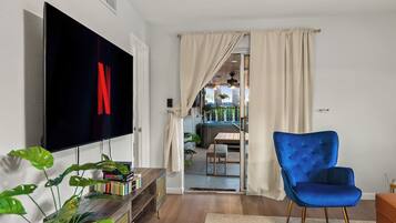A living room with a flat-screen TV, a blue armchair, a bookshelf, and a dining area visible through an open door.