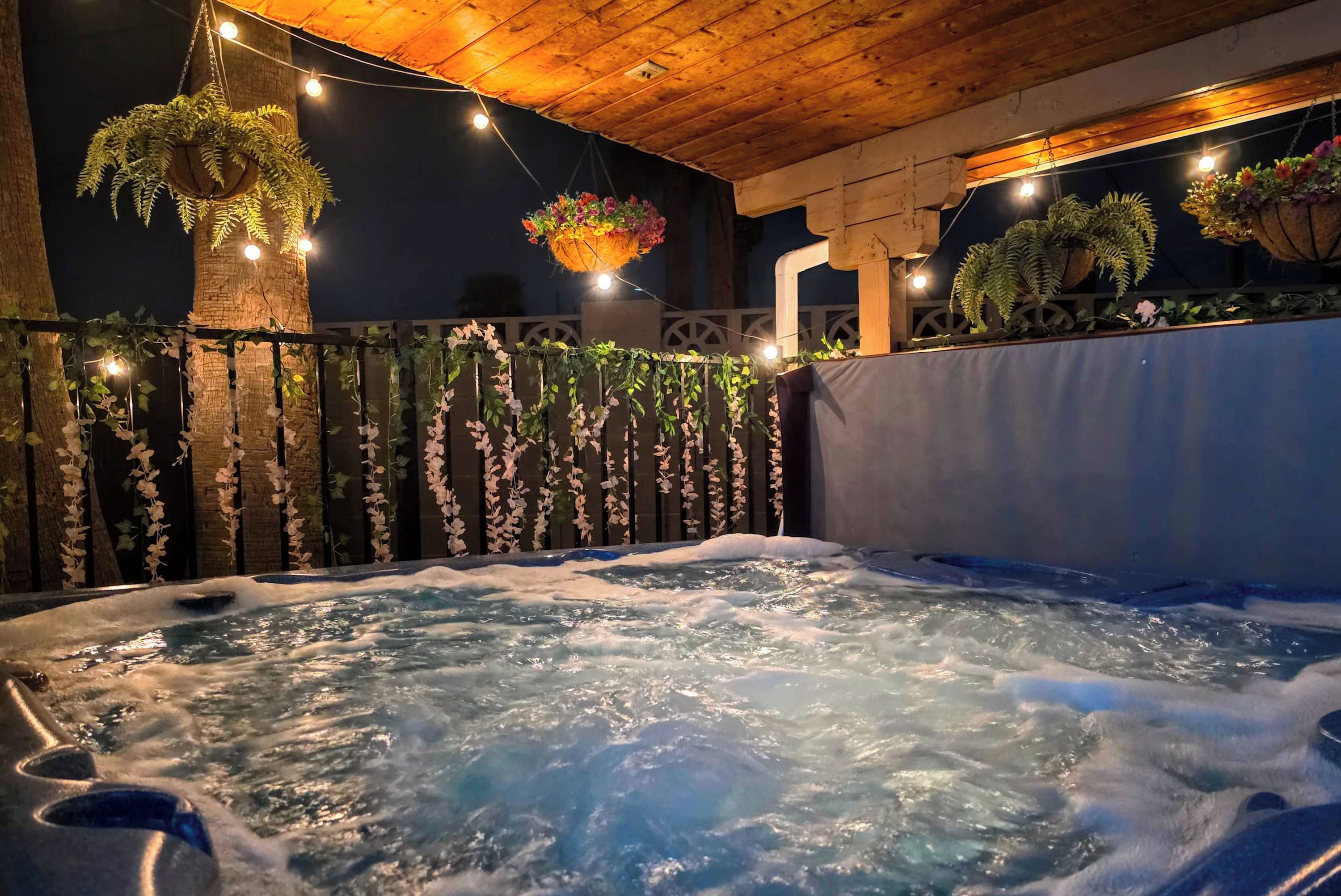 Een hottub met waterstralen, omringd door een houten terras en een hek met hangende planten.