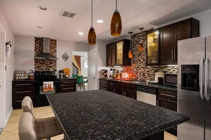 A modern kitchen with a granite countertop, stainless steel appliances, and a dining area with a green chair.
