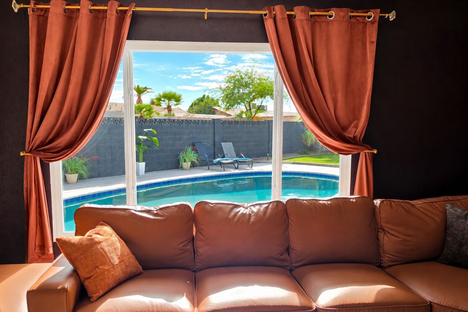 Une pièce avec un canapé en cuir, une fenêtre avec des rideaux et une vue sur une piscine et du mobilier d’extérieur.