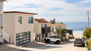 Una calle residencial costera con casas modernas, una camioneta SUV blanca y vista al océano.