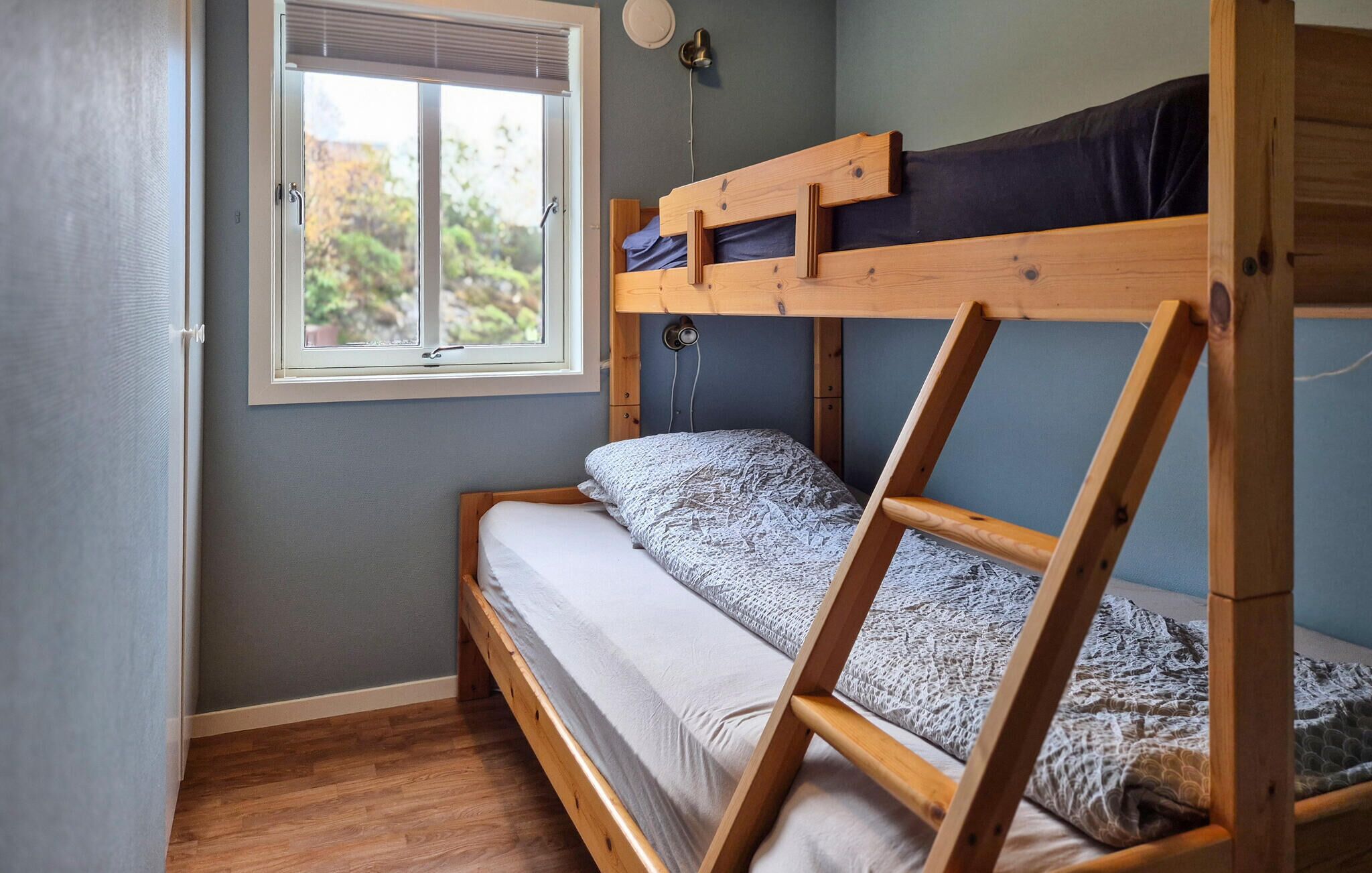 Un lit superposé avec une structure en bois, une fenêtre avec des stores et une lampe fixée au mur.
