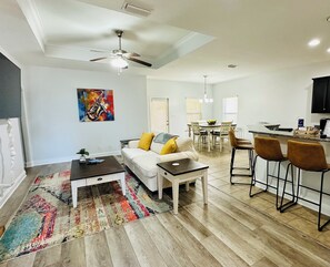 Una sala de estar moderna con un sofá, una mesa de centro y un tapiz de colores. Hay una zona de comedor con una mesa y sillas, y una cocina con barra.