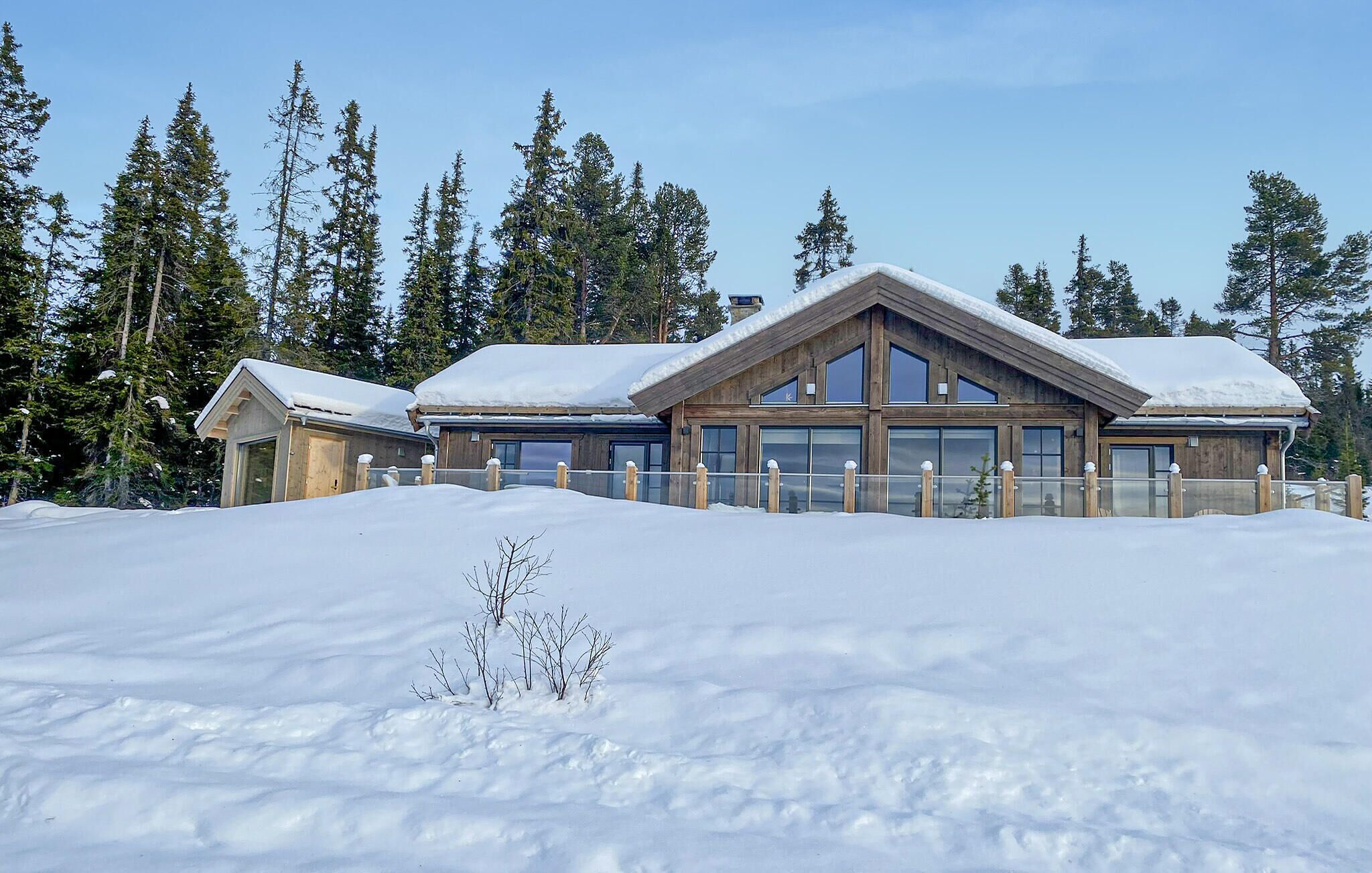 A wooden chalet with large windows and a sloped roof, surrounded by snow and tall pine trees.