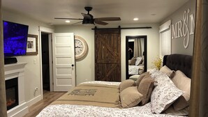 A bedroom with a bed, a television, a fireplace, a ceiling fan, and a large mirror.