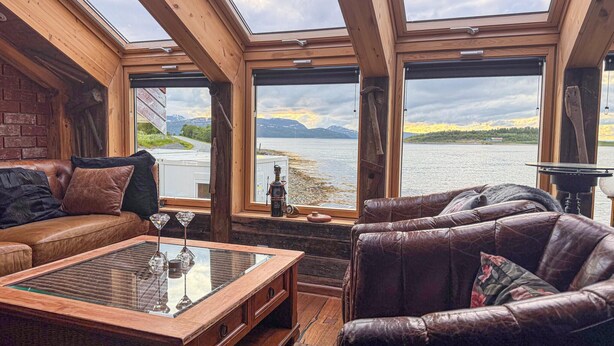 Una habitación acogedora con detalles de madera, sofás de cuero, una mesa de centro de vidrio y grandes ventanales con vistas a un lago y montañas.