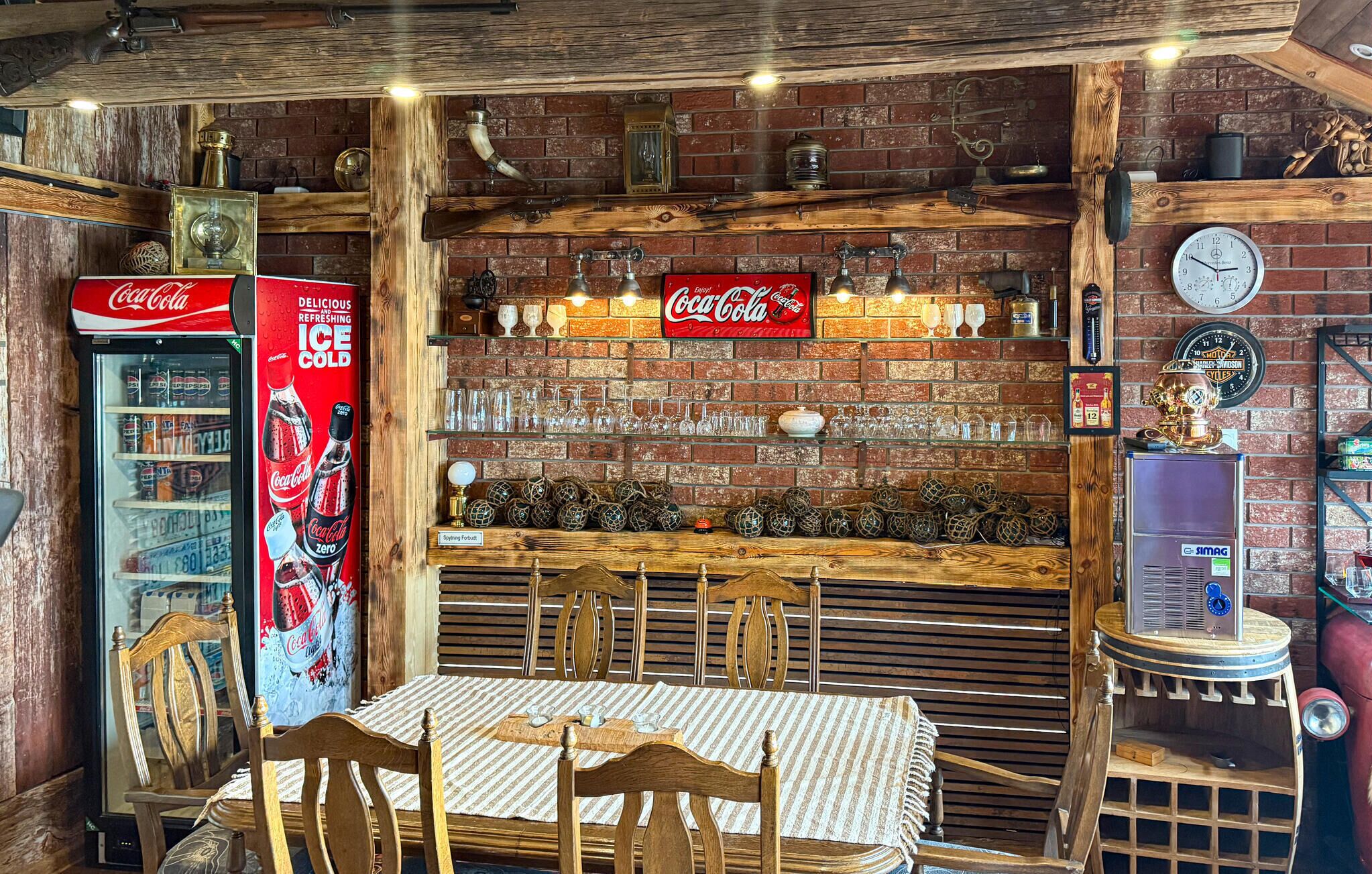 A rustic bar area with a brick wall, vintage Coca-Cola refrigerator, shelves with glasses, and a clock.