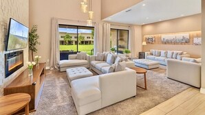 Amplia sala de estar con un televisor grande, varios sofás, una mesa de centro de madera y vistas a un paisaje verde a través de amplios ventanales.