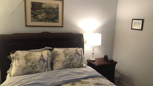 Un lit bien fait, avec une couette aux motifs parisiens, un tableau encadré au-dessus du lit et une table de chevet avec une lampe.
