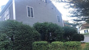 Une maison à deux étages, à l’extérieur gris, entourée d’une végétation dense et d’un mur de pierre.