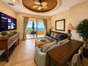 A living room with a large flat-screen TV, a ceiling fan, a dining area with a table and chairs, and a view of the ocean through the sliding glass doors.