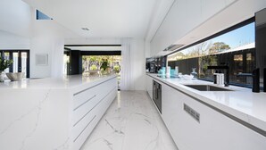 A modern kitchen with white marble countertops, stainless steel appliances, and a large window overlooking a garden. - Villa Capri | Luxe Modern by the Village (Mount Eliza)
