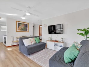 A modern living room with a grey sofa, a dining area, a flat-screen TV, and a ceiling fan. - Tranquil 2-bed Townhouse Close to Beach (Redcliffe)