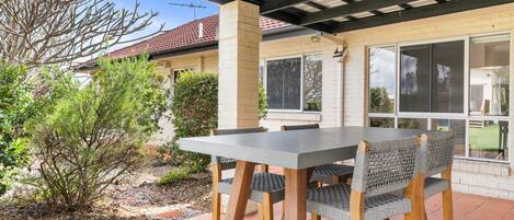 An outdoor patio with a table and chairs, surrounded by a garden and a house with a red-tiled roof.