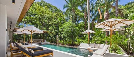 Un área de piscina moderna al aire libre con sillones reclinables y sombrillas, rodeada de exuberante vegetación tropical.