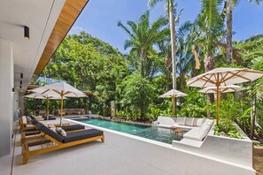 Een zwembad in openlucht met moderne ligbedden en parasols, omgeven door weelderige tropische beplanting.