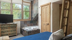 A bedroom with a bed, a dresser, a television, a window, a bathroom with a bathtub, and a wooden door.