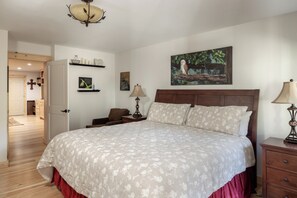 Quarto com cama grande, cabeceira de madeira, mesa de cabeceira com abajur, pequena mesa com abajur, prateleira fixada na parede com objetos decorativos e pintura de uma coruja.